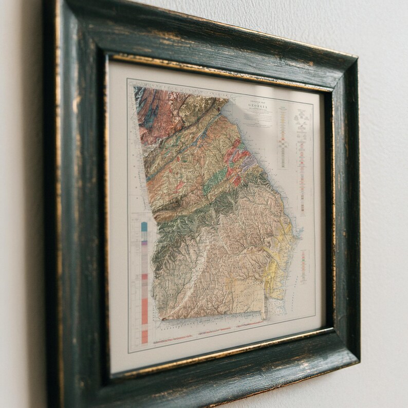 May include: A framed vintage map of Georgia with a detailed topographic map of the state. The map is in shades of green, brown, and yellow, with a color key on the left side. The frame is dark green with gold accents.