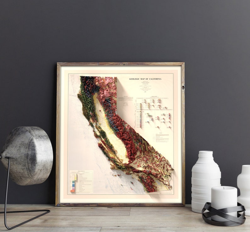 May include: A framed geological map of California, showing a 3D relief of the state's topography. The map uses various colours, including red, green, and yellow, and is set against a neutral backdrop. The frame is made of wood.