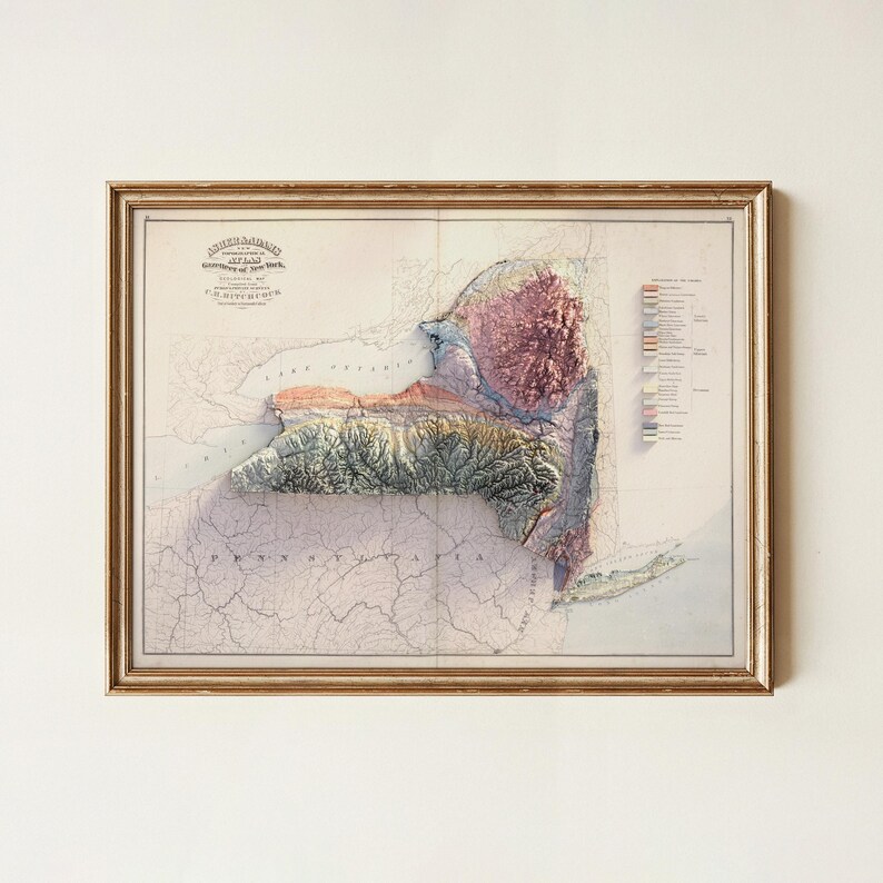 May include: Framed vintage map of New York State with a 3D relief effect, showcasing the topography. The map features shades of green, brown, and purple, with text at the top and a key on the right. The frame is gold.