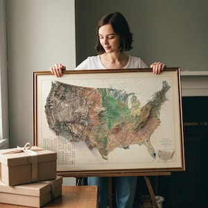 May include: A framed, detailed map of the United States, showcasing topographical features in shades of brown, green, and blue. The map is in a gold-colored frame. The map is held by a person.