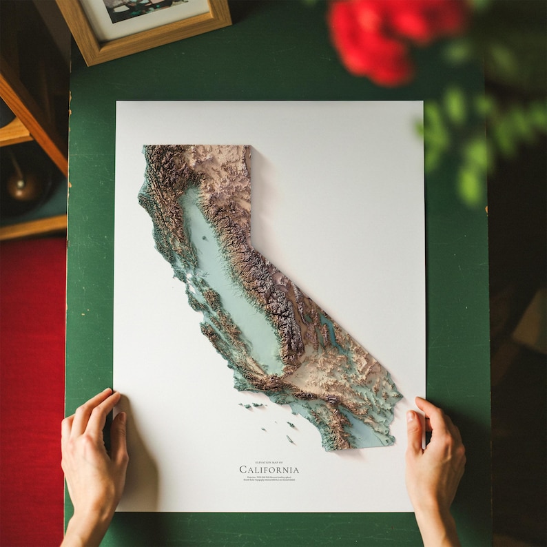 Op de afbeelding: Een gedetailleerde 3D-topografische kaart van Californi&euml;, met een reli&euml;fontwerp in verschillende tinten bruin, beige en blauw. De kaart is gemonteerd op een wit oppervlak, met de naam van de staat onderaan afgedrukt. De kaart wordt door twee handen vastgehouden.