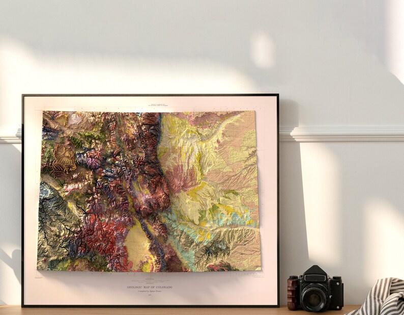 May include: A framed geologic map of Colorado, featuring a detailed relief with various colors representing different geological formations. The map is in a black frame, with the text "GEOLOGIC MAP OF COLORADO" at the bottom. A vintage camera sits nearby.