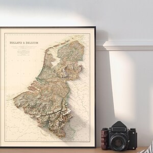 Pode incluir: Um mapa vintage da Holanda e da B&eacute;lgica, mostrando a topografia da regi&atilde;o. O mapa &eacute; impresso em tons de marrom, verde e bege, sobre um fundo branco. O t&iacute;tulo "Holland & Belgium" &eacute; impresso em preto no topo do mapa.
