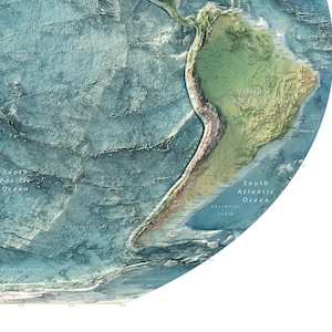 May include: A circular physical map showing the topography of South America and the surrounding ocean floor. The land is rendered in shades of green and brown, while the ocean floor features blue, textured bathymetric relief details.
