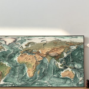 May include: A framed world map with a raised relief design, showing the continents and oceans in shades of green, brown, and blue. The map is framed in black and is hanging on a white wall.