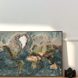 May include: A framed world map with a raised relief design, showing the continents and oceans in shades of blue, green, brown, and white. The map is framed in black and is hanging on a white wall.