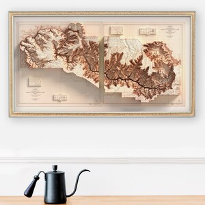May include: Framed relief map of the Grand Canyon National Park, showcasing the topography in shades of brown and beige. The map is encased in a gold-colored frame. A black gooseneck kettle with a wooden handle sits on a wooden surface.