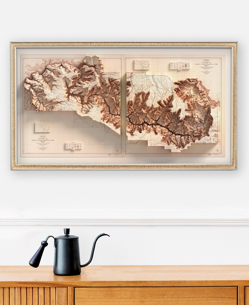 May include: Framed relief map of the Grand Canyon National Park, showcasing the topography in shades of brown and beige. The map is encased in a gold-colored frame. A black gooseneck kettle with a wooden handle sits on a wooden surface.