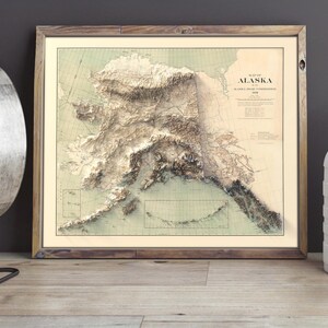 Large-format Alaska topographic map in a distressed barnwood frame, styled above a quilted bed in a rustic lodge bedroom. The artwork highlights the Brooks Range and Arctic coastline, paired with a lantern, leather-bound journal, and pinecones—a dreamy gift for cabin owners, hikers, or anyone who cherishes the quiet grandeur of Alaska’s remote landscapes