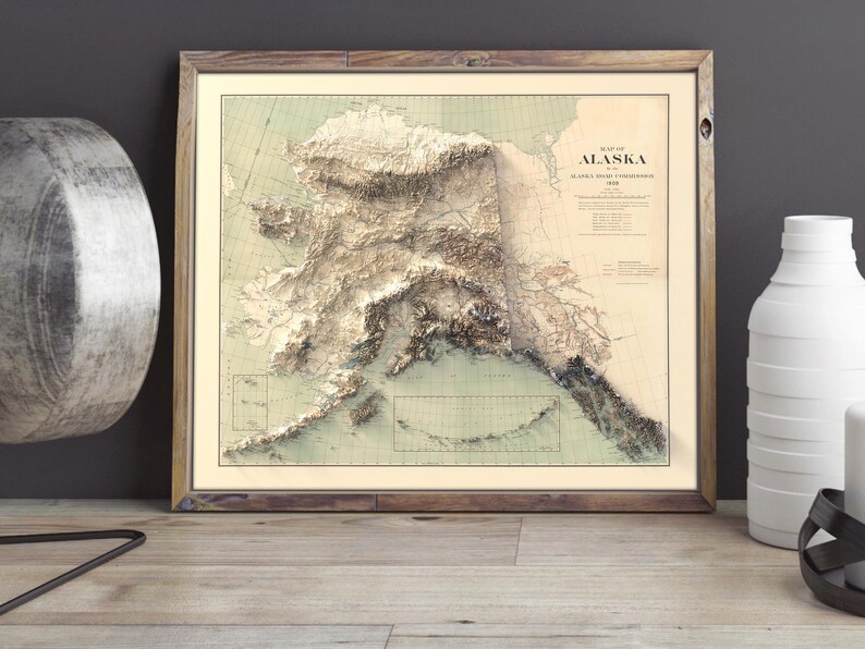 Large-format Alaska topographic map in a distressed barnwood frame, styled above a quilted bed in a rustic lodge bedroom. The artwork highlights the Brooks Range and Arctic coastline, paired with a lantern, leather-bound journal, and pinecones—a dreamy gift for cabin owners, hikers, or anyone who cherishes the quiet grandeur of Alaska’s remote landscapes