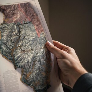 May include: A detailed relief map of Illinois, showcasing the state's topography with varying shades of brown, gray, and red. The map is held by a person, revealing the intricate details of the terrain. The map appears to be a print.