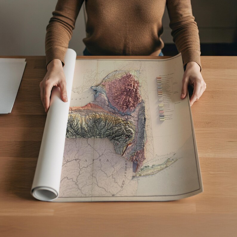 May include: A rolled-up map of New York State is being unrolled on a wooden surface. The map features a 3D topographic design with shades of green, brown, and pink, and includes detailed geographical features. The map is printed on aged paper.