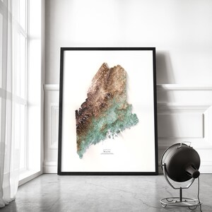 May include: A framed print of a topographic map of Maine, featuring a 3D-like design with shades of brown, green, and blue. The print is in a black frame and is hanging on a white wall.
