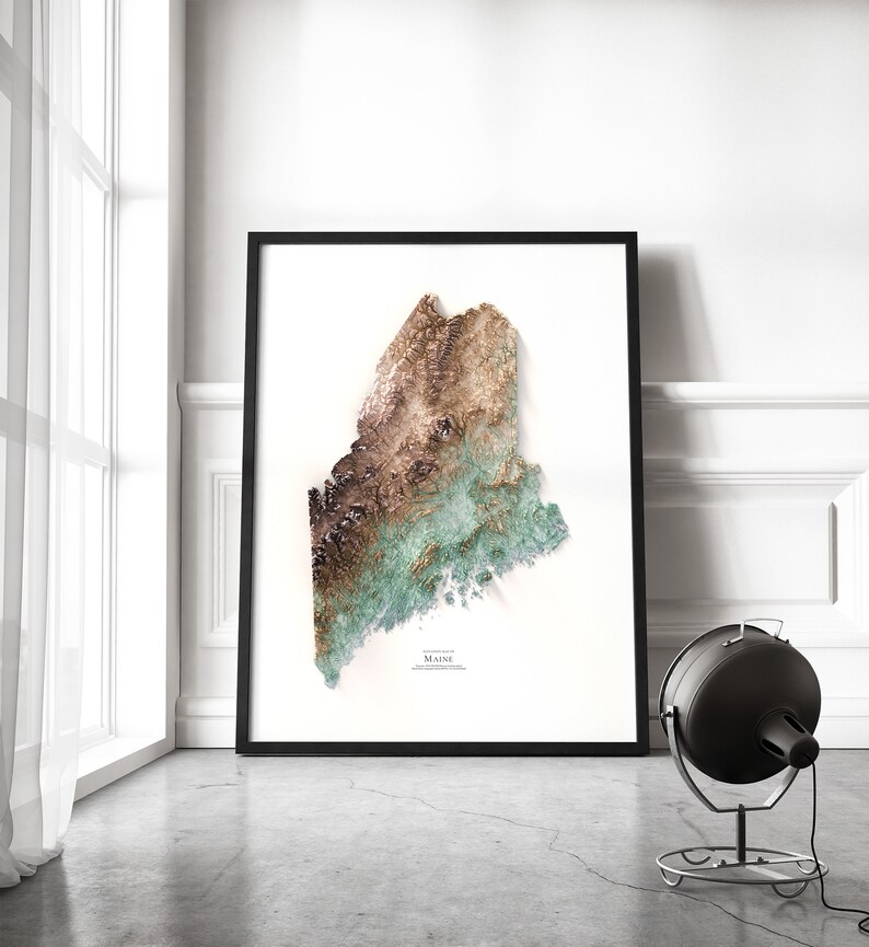 May include: A framed print of a topographic map of Maine, featuring a 3D-like design with shades of brown, green, and blue. The print is in a black frame and is hanging on a white wall.