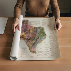 Op de afbeelding: Een gedetailleerde, veelkleurige kaart van Texas, met topografische kenmerken, wordt uitgerold op een houten oppervlak. De kaart benadrukt het diverse landschap van de staat, inclusief de kustlijn en de binnenlandse regio's. De kaart is gedrukt op cr&egrave;mekleurig papier.
