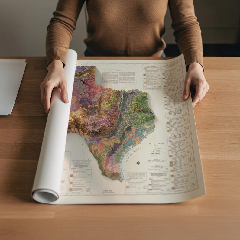 Op de afbeelding: Een gedetailleerde, veelkleurige kaart van Texas, met topografische kenmerken, wordt uitgerold op een houten oppervlak. De kaart benadrukt het diverse landschap van de staat, inclusief de kustlijn en de binnenlandse regio's. De kaart is gedrukt op cr&egrave;mekleurig papier.