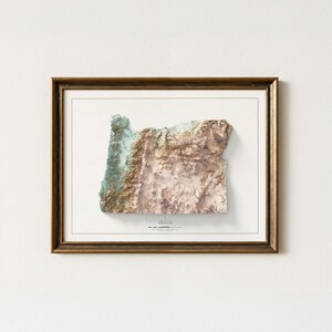 Puede incluir: Mapa topogr&aacute;fico 3D enmarcado de Oreg&oacute;n, con un relieve detallado en tonos marrones, verdes y beige. El mapa est&aacute; sobre un fondo blanco y encerrado en un marco de madera marr&oacute;n, ofreciendo una representaci&oacute;n texturizada y art&iacute;stica del paisaje del estado.