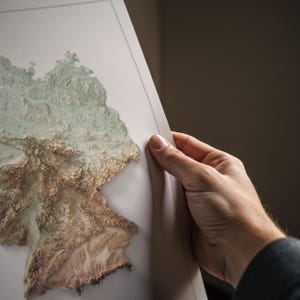 May include: A detailed relief map of Germany, showing its topography in shades of brown and green. The map is held by a person, highlighting the intricate details of the landscape. The map appears to be a framed print.
