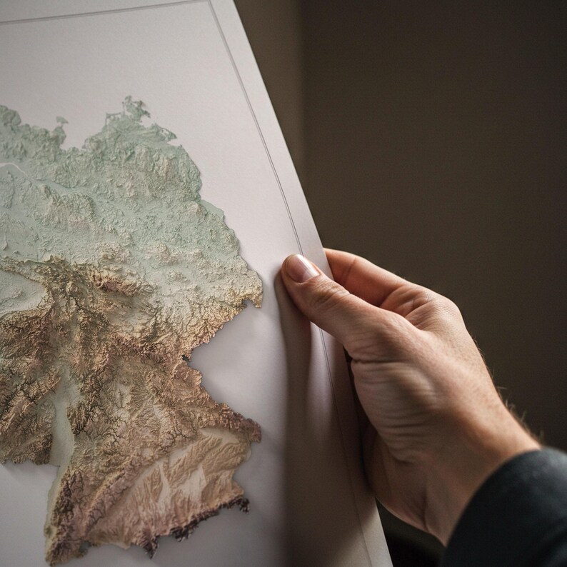 May include: A detailed relief map of Germany, showing its topography in shades of brown and green. The map is held by a person, highlighting the intricate details of the landscape. The map appears to be a framed print.