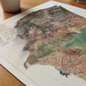 May include: A detailed, three-dimensional map print with a relief effect, showcasing a region with varying elevations. The map features a color palette of earthy tones, including browns, greens, and tans, with a white border. The print is displayed on a wooden surface.