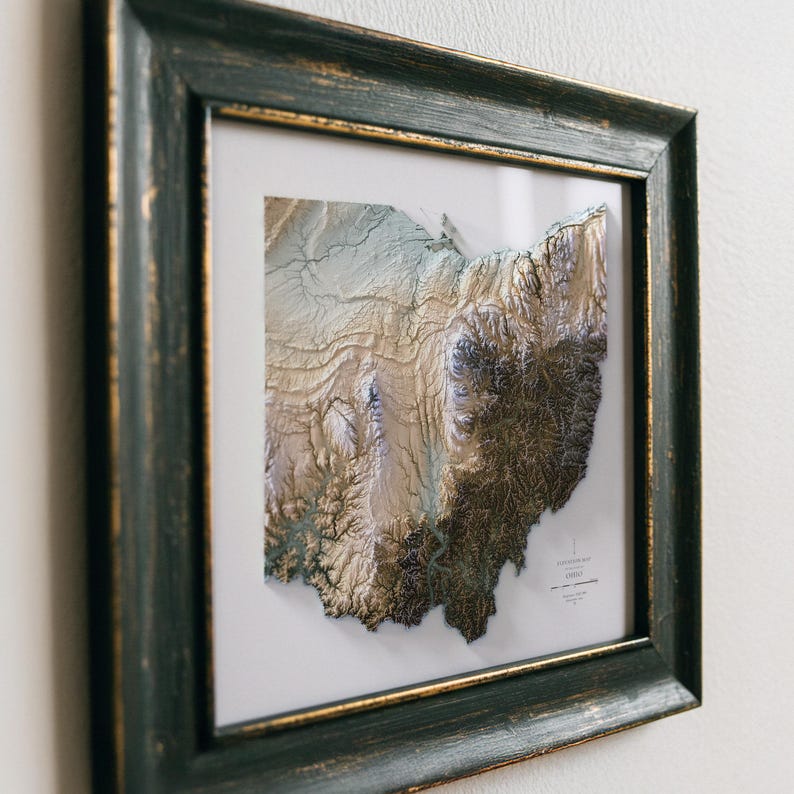 K&ouml;nnte beinhalten: Gerahmte 3D-Topografische Karte von Ohio mit detaillierten Reliefs und Farbvariationen. Die Karte ist in einem verwitterten, dunkelgr&uuml;nen Holzrahmen mit goldenem Rand eingefasst, was eine Vintage-&Auml;sthetik erzeugt. Die Karte befindet sich vor einem wei&szlig;en Hintergrund.