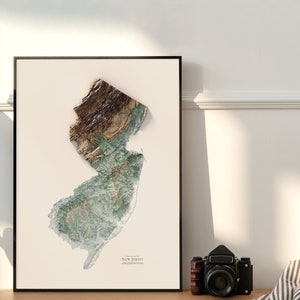 May include: A framed print of a topographic map of New Jersey. The map is in shades of green, brown, and white, and shows the state's terrain in detail. The print is framed in a black frame and has a white mat.