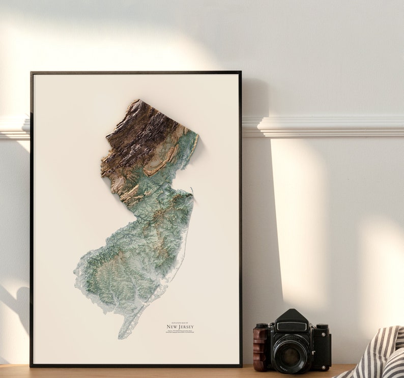 May include: A framed print of a topographic map of New Jersey. The map is in shades of green, brown, and white, and shows the state's terrain in detail. The print is framed in a black frame and has a white mat.