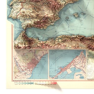 May include: A vintage map of Spain with a detailed inset map of Barcelona. The map is printed on a cream-colored paper with blue, green, and brown lines and text. The map shows the coastline, mountains, and cities of Spain.
