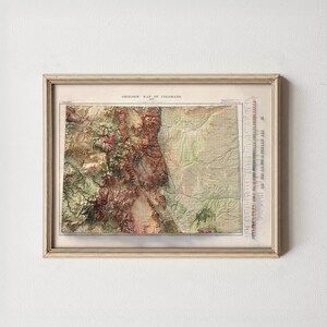 May include: Framed vintage geologic map of Colorado. The map features a detailed relief with shades of green, brown, and red, encased in a cream-colored wooden frame. The map includes text at the top and a scale along the right edge.