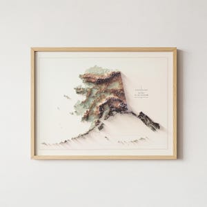 May include: A framed 3D elevation map of Alaska, showcasing the state's topography in shades of green, brown, and beige. The map is set against a cream-colored background and framed in light wood. The text "Elevation Map Alaska" is visible.