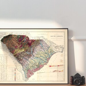 Pode incluir: Um mapa vintage da Carolina do Sul com um efeito de relevo 3D. O mapa &eacute; impresso em tons de marrom, verde e azul, e mostra a topografia do estado. O mapa &eacute; emoldurado em preto e est&aacute; pendurado em uma parede branca.