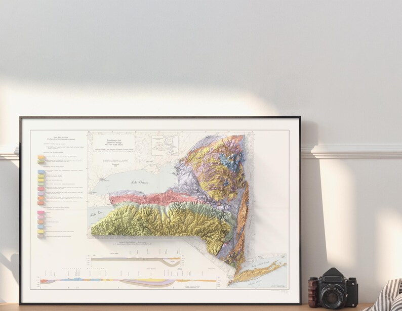 Op de afbeelding: Een ingelijste 3D-kaart van de staat New York, met topografische kenmerken in verschillende kleuren. De kaart zit in een zwart frame, met een vintage camera op een houten oppervlak. De kaart bevat tekst en een sleutel.