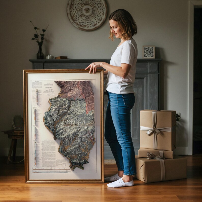 May include: A framed map of Illinois with a detailed relief, showcasing the state's topography in shades of brown, gray, and green. The map is held by a person, and the frame is gold-colored. The map includes text and is displayed indoors.