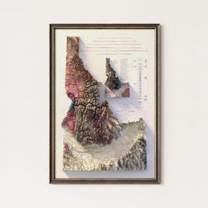 May include: Framed 3D relief map of Idaho, featuring detailed topographical features in shades of brown, pink, and green. The map is encased in a dark wooden frame, showcasing the state's landscape with precision and artistic flair.