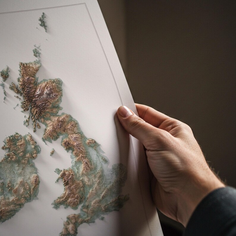 May include: A detailed relief map of Great Britain and Ireland, featuring a 3D topographic design. The map is rendered in shades of brown and green, with a white border. The map is held by a person's hand.