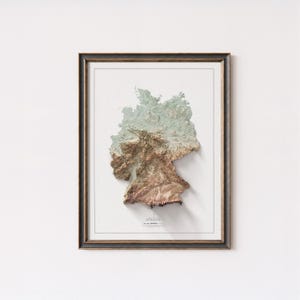 May include: A framed 3D relief map of Germany, depicting the country's topography in green, brown, and beige hues. The map is set against a white background, within a dark wooden frame with a gold inner border. The word "GERMANY" is printed at the bottom.
