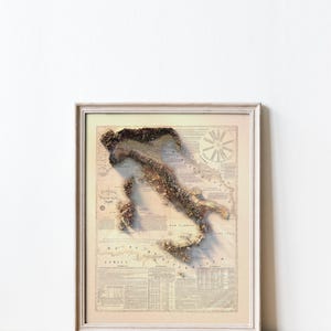 May include: A vintage-style map of Italy in a white frame. The map is a 3D relief map with a beige background and brown and green topographical features. The map is surrounded by text and a compass rose.