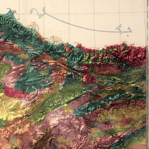 May include: A detailed topographic map of a mountainous region with various shades of green, red, and yellow representing different elevations. The map features a coastline and a few Arabic script labels.