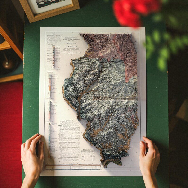May include: A detailed relief map of Illinois, showcasing the state's topography with varying shades of brown, green, and blue. The map is printed on a white paper and held by two hands. The map includes the text "ILLINOIS".