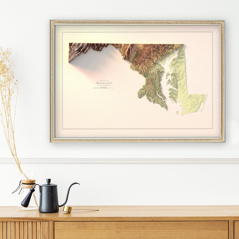 May include: A framed map of Maryland with a raised relief design. The map is printed in shades of green, brown, and beige, with a white background. The state name "MARYLAND" is printed in black at the bottom of the map.