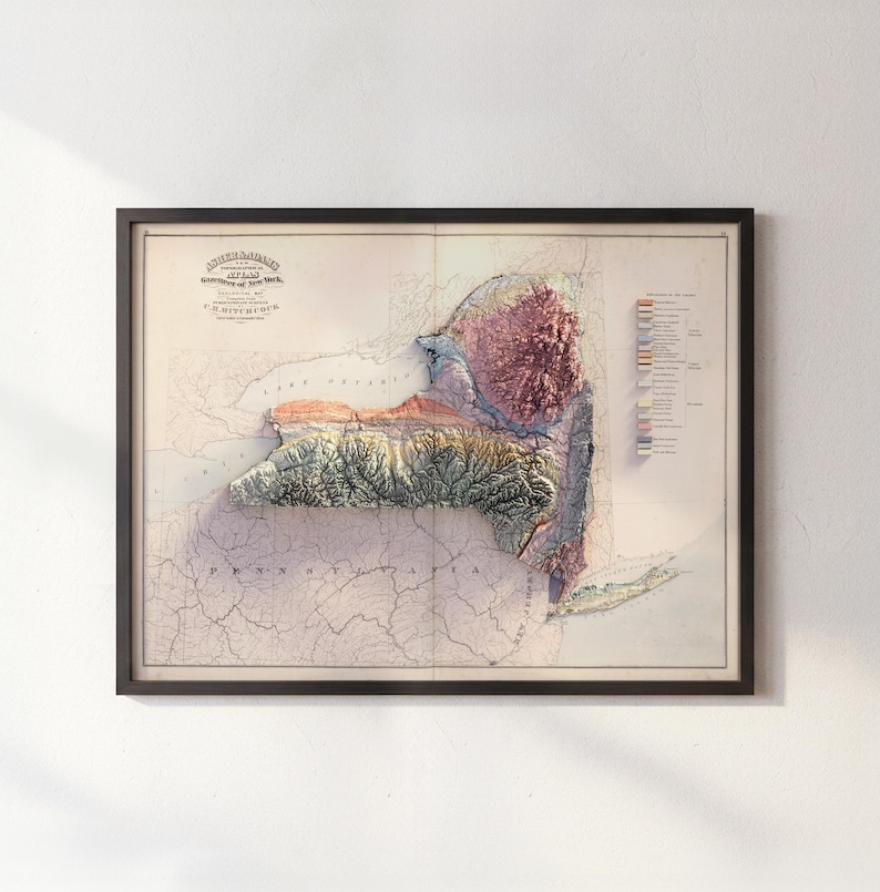 May include: Framed vintage map of New York State, featuring a 3D topographic relief. The map displays a detailed view of the state's terrain, with colors indicating elevation changes. The frame is black, and the map includes text at the top.
