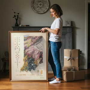 May include: A framed map of Indiana with a detailed topographical design. The map features a variety of colours, including shades of green, purple, and brown, and is encased in a gold-coloured frame. The map is held by a person.
