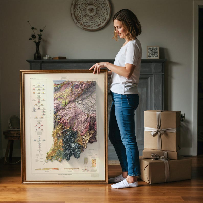 May include: A framed map of Indiana with a detailed topographical design. The map features a variety of colours, including shades of green, purple, and brown, and is encased in a gold-coloured frame. The map is held by a person.