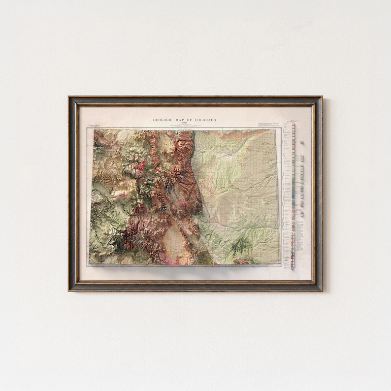 May include: Framed vintage map of Colorado, showcasing a detailed geological representation. The map features a color palette of greens, browns, and reds, with the title "GEOLOGY MAP OF COLORADO" at the top. The map is encased in a dark brown wooden frame.