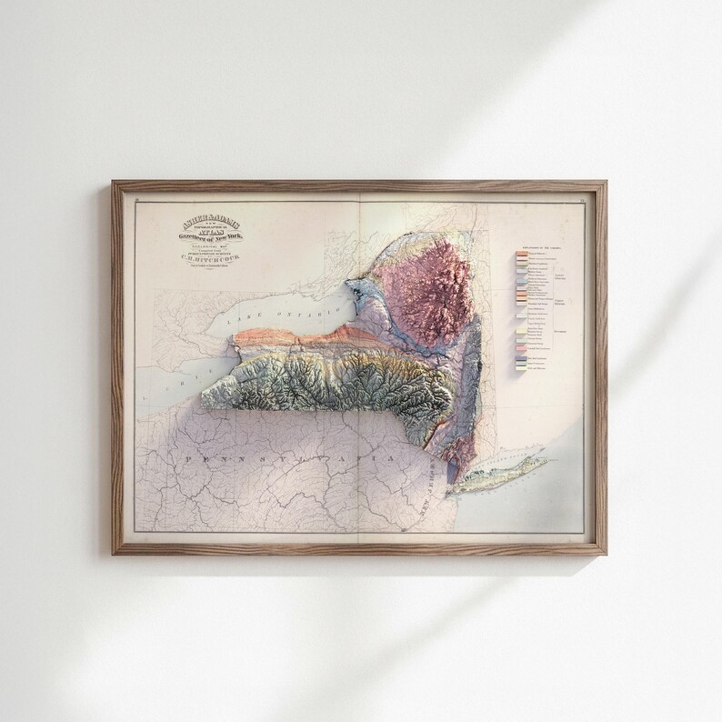 May include: Framed vintage map of New York State, featuring a 3D relief effect. The map displays detailed topography with shades of green, brown, and pink, and includes text at the top left corner. The map is encased in a light brown wooden frame.