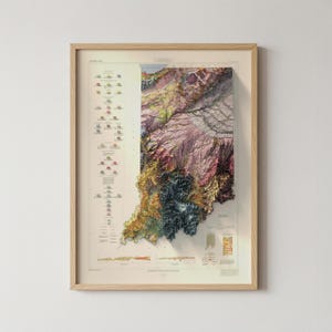 May include: Framed topographical map of Indiana, with a detailed 3D rendering of the state's terrain. The map uses various colours, including greens, browns, and purples, with a key and additional data on the left. The frame is made of light-coloured wood.