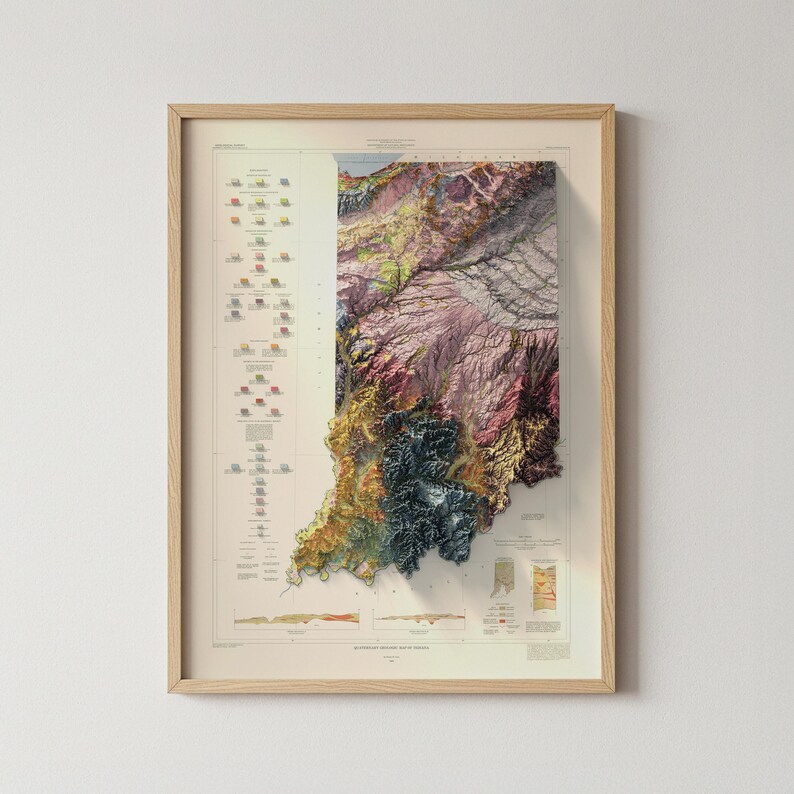 May include: Framed topographical map of Indiana, with a detailed 3D rendering of the state's terrain. The map uses various colours, including greens, browns, and purples, with a key and additional data on the left. The frame is made of light-coloured wood.