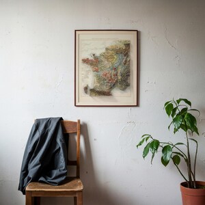 May include: A framed vintage map of France, featuring detailed topographical information and a brown wooden frame. The map is mounted on a white wall, with a wooden chair and a dark jacket draped over it. A potted plant is in the corner.