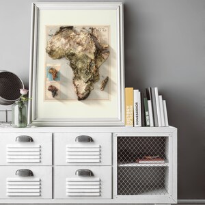 May include: A white cabinet with four drawers and a wire shelf. The cabinet has a framed map of Africa hanging above it. The map is in a white frame and has a vintage look. There are books on top of the cabinet, including one titled "History of Design".