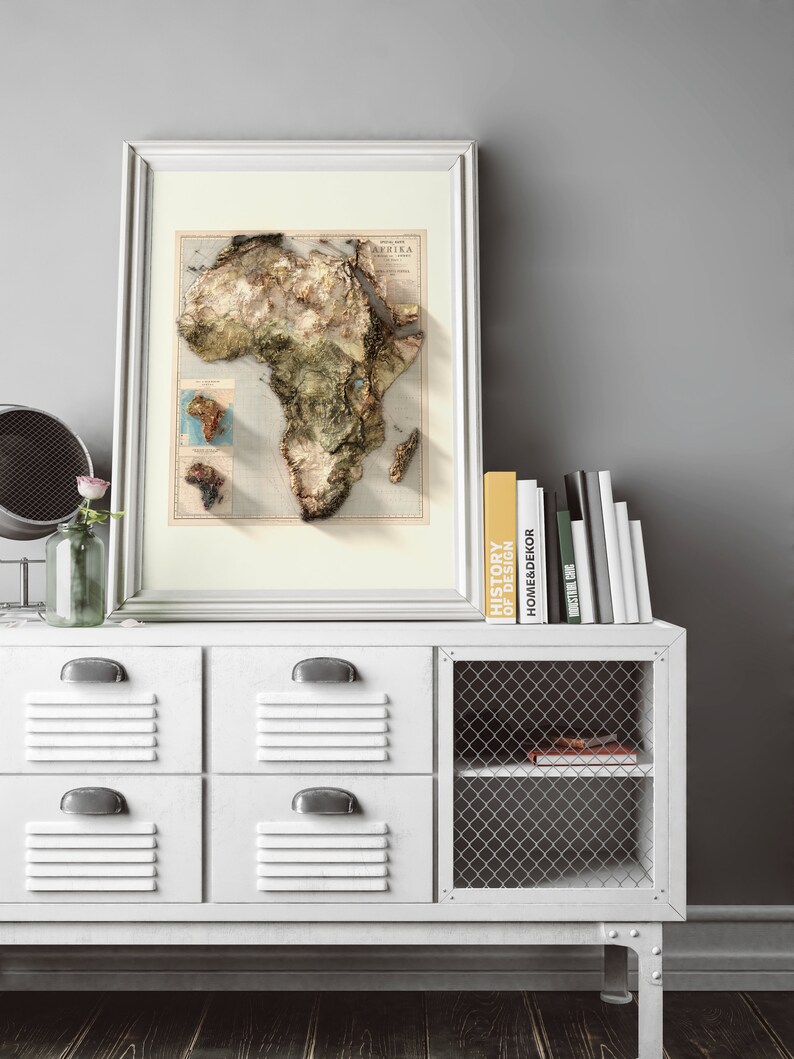 May include: A white cabinet with four drawers and a wire shelf. The cabinet has a framed map of Africa hanging above it. The map is in a white frame and has a vintage look. There are books on top of the cabinet, including one titled "History of Design".